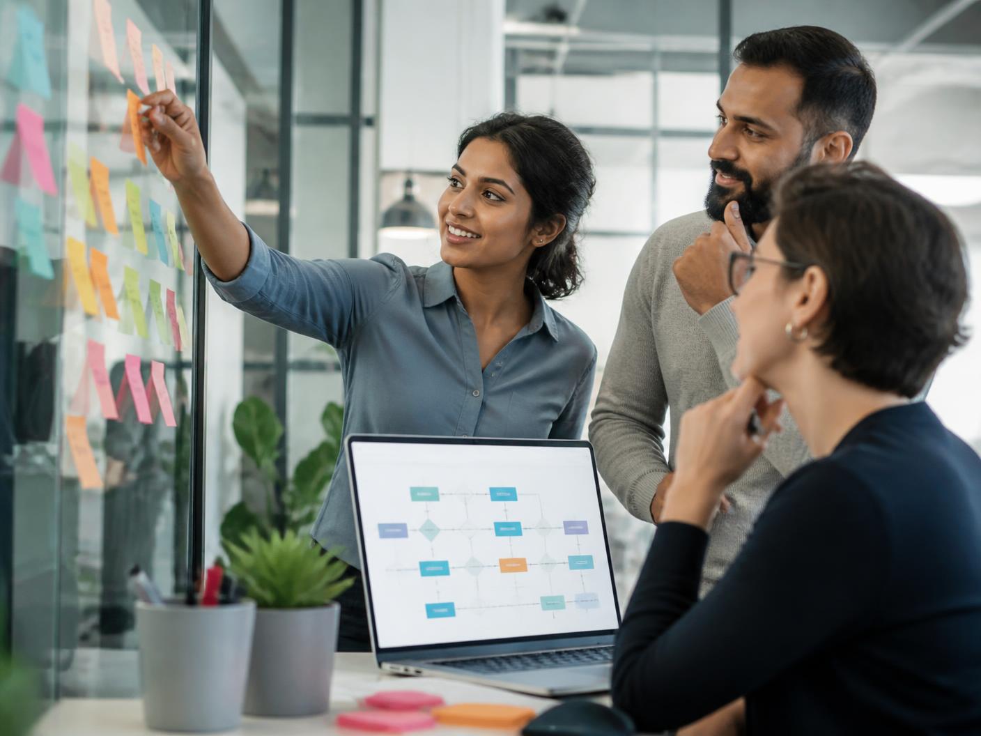 A small team of working professionals mapping an automation workflow on a glass wall beside a laptop showing an agent flowchart